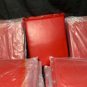 May include: Several red rectangular items, possibly cushions or pads, are visible. One is fully exposed, showing a smooth, shiny surface with grommets along the top edge. The others are wrapped in clear plastic. The background is a dark, solid color.