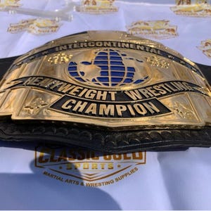 May include: A gold-colored Intercontinental Heavyweight Wrestling Champion belt. The belt features a blue globe design and the words "Heavyweight Wrestling Champion" in black lettering. The belt is on a white surface.