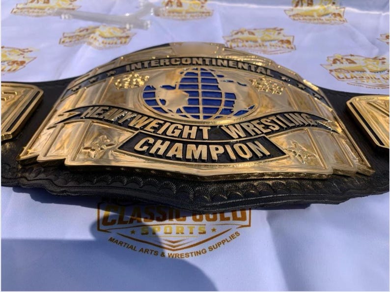 May include: A gold-colored Intercontinental Heavyweight Wrestling Champion belt. The belt features a blue globe design and the words "Heavyweight Wrestling Champion" in black lettering. The belt is on a white surface.