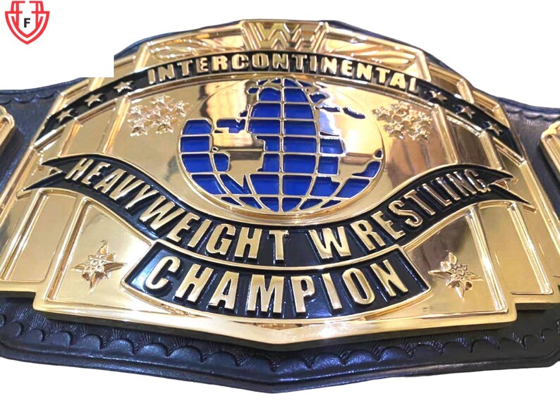 May include: A gold-colored wrestling championship belt with black and blue accents. The belt features the words "Intercontinental", "Heavyweight Wrestling Champion", and a blue globe design. The belt is made of leather and metal.
