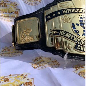 May include: A close-up of a wrestling championship belt. The belt features a black leather strap with gold-colored metal plates. The plates display the words "INTERCONTINENTAL HEAVYWEIGHT CHAMPION" and a globe design. The background is white with gold logos.