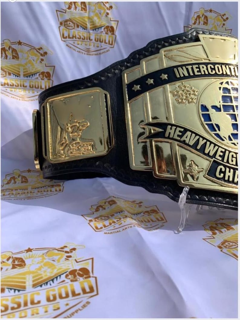 May include: A close-up of a wrestling championship belt. The belt features a black leather strap with gold-colored metal plates. The plates display the words "INTERCONTINENTAL HEAVYWEIGHT CHAMPION" and a globe design. The background is white with gold logos.