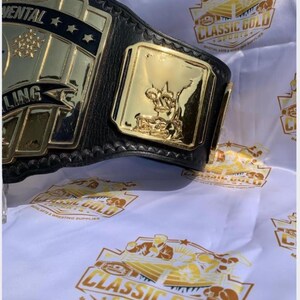 May include: A gold and black wrestling championship belt with the word "Continental" and stars on the main plate. The belt has a gold side plate with a figure. The background features a white cloth with the "Classic Gold Sports" logo.