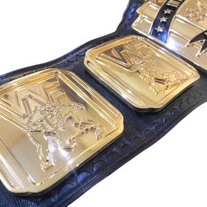 May include: Close-up of a gold and black wrestling championship belt. The belt features gold plates with the letters "WWE" and wrestler figures. The belt is made of black leather and has blue and white accents.