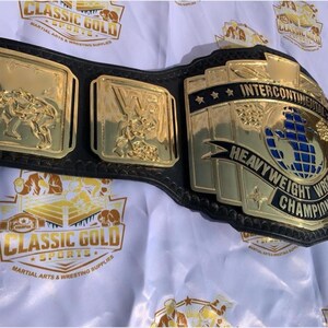 May include: A gold and black wrestling championship belt. The belt features gold plates with wrestling figures and the words "Intercontinental Heavyweight Wrestling Champion". The belt is on a white background with the words "Classic Gold Sports" printed on it.