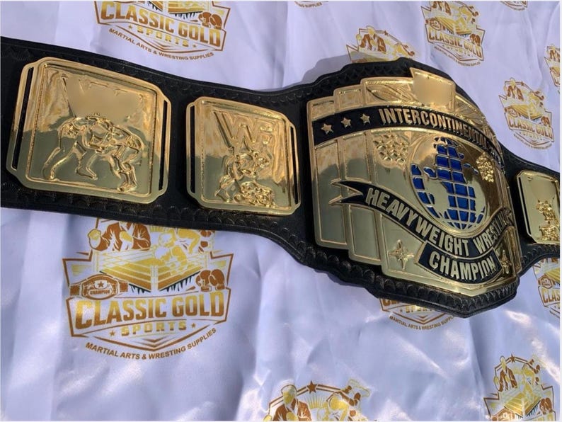 May include: A gold and black wrestling championship belt. The belt features gold plates with wrestling figures and the words "Intercontinental Heavyweight Wrestling Champion". The belt is on a white background with the words "Classic Gold Sports" printed on it.