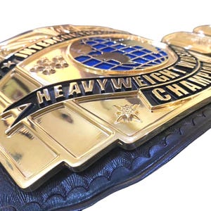 May include: Close-up of a gold and black heavyweight championship belt. The belt features the words "International Heavyweight Champion" and a blue globe design. The belt is made of gold-colored metal and black leather.