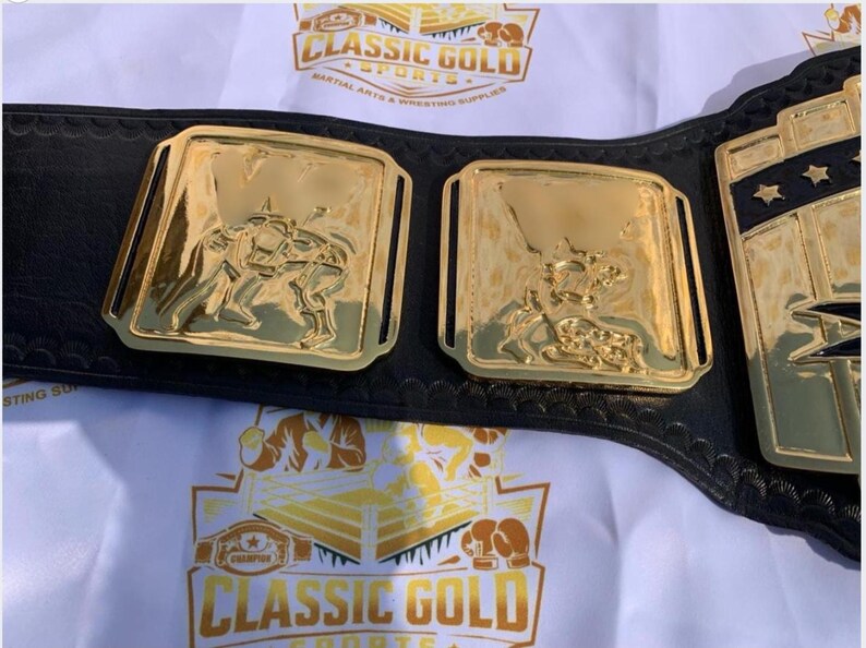 May include: A gold-colored wrestling championship belt with a black leather strap. The belt features gold rectangular plates with detailed relief images of wrestlers. The text "CLASSIC GOLD SPORTS" is visible in the background.