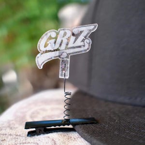 May include: A clear acrylic hat pin with the word "GRZ" in white lettering. The pin is attached to a black metal clip with a small spring. The pin is attached to a black baseball cap.