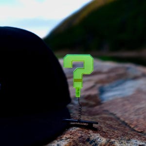 May include: A black baseball cap with a large number 3 embroidered in light blue on the side. A neon green question mark pin is attached to the cap, with a black wire and spring.