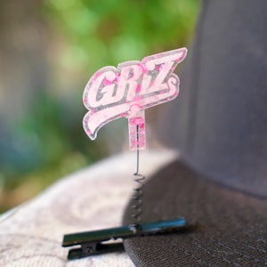 Puede incluir: Un pin con el logotipo "GRIZ" en rosa y blanco sujeto a un sombrero negro con un clip. El pin tiene una textura brillante y está conectado a un clip negro mediante un resorte metálico. El sombrero tiene un diseño circular blanco.