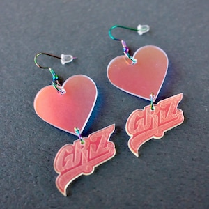 May include: A pair of heart-shaped earrings with the word "GIRLZ" in a script font. The earrings are a gradient of pink and peach with iridescent hooks.