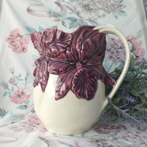 May include: A white ceramic pitcher with a purple floral design. The pitcher has a wide mouth and a handle. The floral design is raised and detailed.