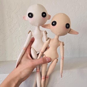 May include: Two handmade fabric dolls with button eyes and small, pointed ears. One doll is white, the other is tan. Both dolls have articulated limbs and are held together by a hand.