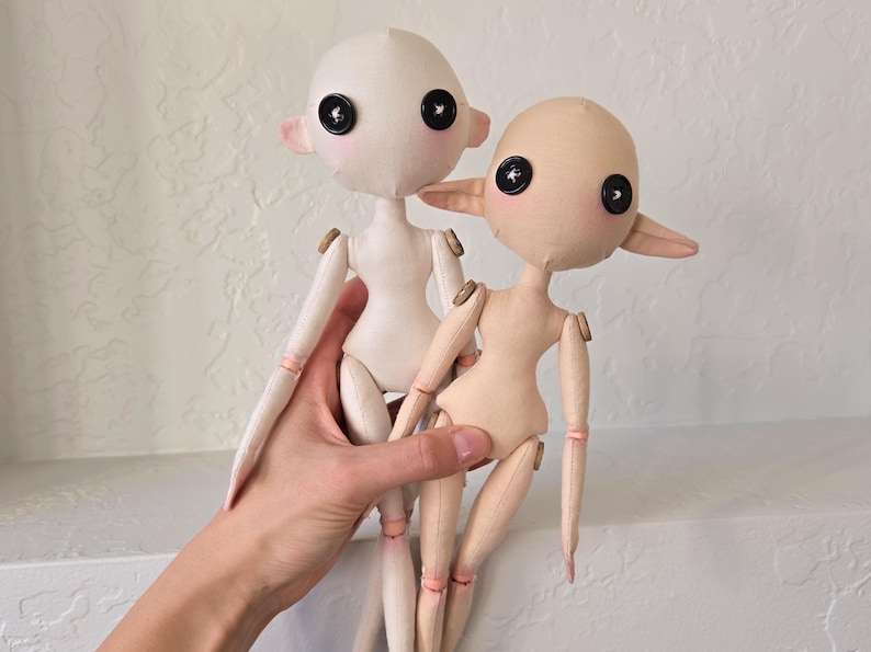 May include: Two handmade fabric dolls with button eyes and small, pointed ears. One doll is white, the other is tan. Both dolls have articulated limbs and are held together by a hand.