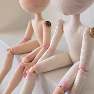 May include: Two handmade cloth dolls with light skin tones are seated side-by-side. The dolls have button joints and painted facial features. Their limbs are accented with pink paint. The dolls are resting on a white surface.