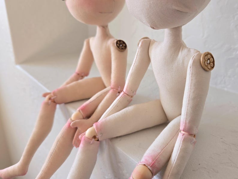 May include: Two handmade cloth dolls with light skin tones are seated side-by-side. The dolls have button joints and painted facial features. Their limbs are accented with pink paint. The dolls are resting on a white surface.