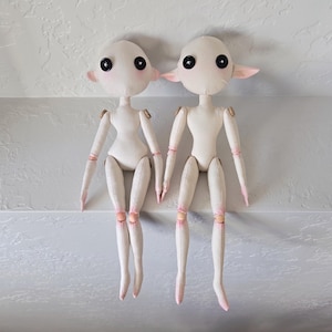 May include: Two handmade fabric dolls with pale skin, large black eyes, and pink accents on their ears, hands, and feet. The dolls have jointed arms and legs, and are sitting side-by-side. The dolls are a unique, whimsical craft.