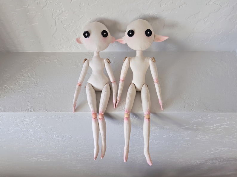 May include: Two handmade fabric dolls with pale skin, large black eyes, and pink accents on their ears, hands, and feet. The dolls have jointed arms and legs, and are sitting side-by-side. The dolls are a unique, whimsical craft.