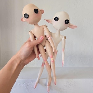 May include: Two handmade fabric dolls with button eyes and rosy cheeks. The dolls have articulated limbs and pointed ears. One doll is held in a hand, and the other stands behind it. The dolls are primarily beige and white with pink accents.