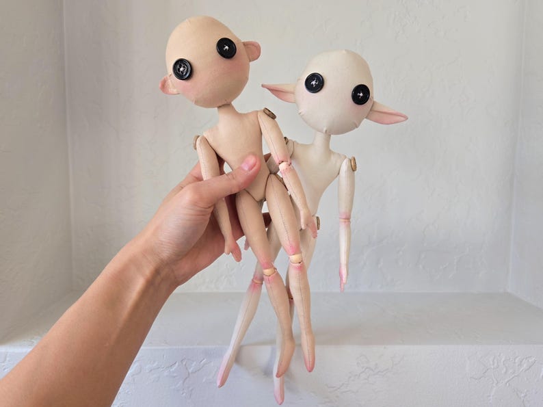 May include: Two handmade fabric dolls with button eyes and rosy cheeks. The dolls have articulated limbs and pointed ears. One doll is held in a hand, and the other stands behind it. The dolls are primarily beige and white with pink accents.