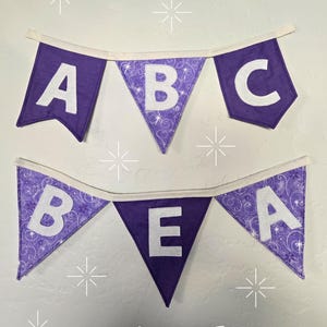K&ouml;nnte beinhalten: Zwei lila Stoffbanner mit wei&szlig;en Buchstaben, die "ABC" und "BEA" buchstabieren. Die Banner sind mit wei&szlig;em Stoff beschnitten und h&auml;ngen an einer wei&szlig;en Wand. Das Wort "triangle" ist unten aufgedruckt.