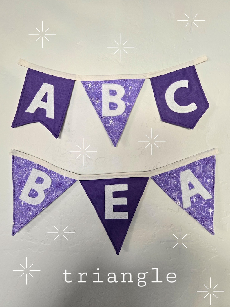 K&ouml;nnte beinhalten: Zwei lila Stoffbanner mit wei&szlig;en Buchstaben, die "ABC" und "BEA" buchstabieren. Die Banner sind mit wei&szlig;em Stoff beschnitten und h&auml;ngen an einer wei&szlig;en Wand. Das Wort "triangle" ist unten aufgedruckt.