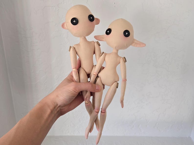 May include: Two handmade cloth dolls with button eyes and blush cheeks. The dolls have jointed wooden limbs and are dressed in a nude color. The dolls are held in a person's hand.