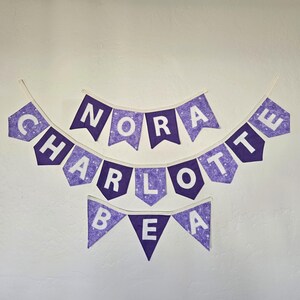 K&ouml;nnte beinhalten: Drei lila Stoffbanner mit wei&szlig;en Buchstaben, die "NORA", "CHARLOTTE" und "BEA" buchstabieren. Die Banner sind mit einem Wirbelmuster verziert und h&auml;ngen an einer wei&szlig;en Wand.