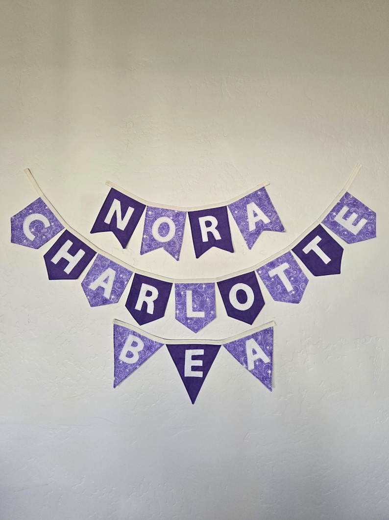 K&ouml;nnte beinhalten: Drei lila Stoffbanner mit wei&szlig;en Buchstaben, die "NORA", "CHARLOTTE" und "BEA" buchstabieren. Die Banner sind mit einem Wirbelmuster verziert und h&auml;ngen an einer wei&szlig;en Wand.