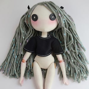 May include: A handmade fabric doll with pale skin, black button eyes, and pink cheeks. The doll has long, textured sage green yarn hair with small colorful flecks and wears a short-sleeved black crop top. The arms and legs are articulated.