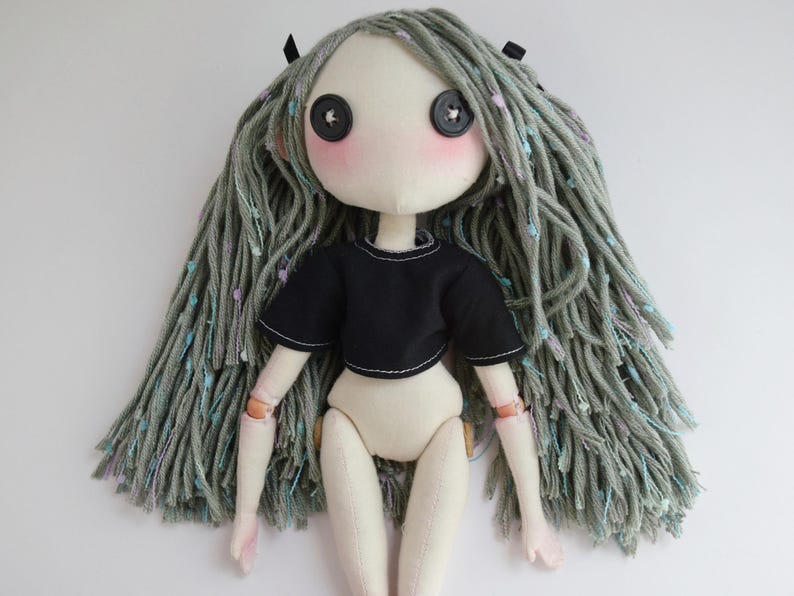 May include: A handmade fabric doll with pale skin, black button eyes, and pink cheeks. The doll has long, textured sage green yarn hair with small colorful flecks and wears a short-sleeved black crop top. The arms and legs are articulated.