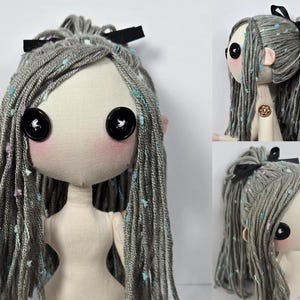 May include: A handmade doll with a fabric face, button eyes, and gray yarn hair adorned with a black bow. The doll has pink cheeks and ears, and its hair is speckled with colorful accents. The doll's body is made of a light-colored fabric.