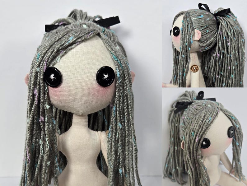 May include: A handmade doll with a fabric face, button eyes, and gray yarn hair adorned with a black bow. The doll has pink cheeks and ears, and its hair is speckled with colorful accents. The doll's body is made of a light-colored fabric.