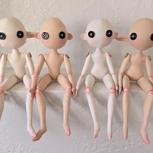 May include: Four handmade cloth dolls with button eyes and pointed ears. The dolls have articulated limbs and are in various skin tones, with pink accents on the knees and elbows. They are sitting on a white surface.