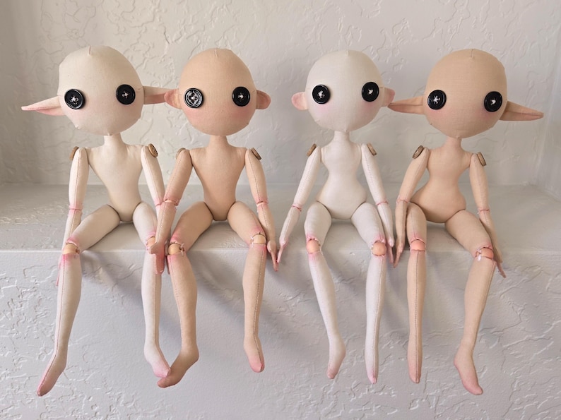 May include: Four handmade cloth dolls with button eyes and pointed ears. The dolls have articulated limbs and are in various skin tones, with pink accents on the knees and elbows. They are sitting on a white surface.