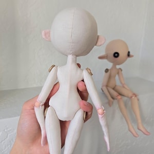 May include: Two handmade doll figures. One doll is held in a hand, showing the back of the doll with a round head, small ears, and jointed limbs. The other doll is seated, with a similar design and a light skin tone.
