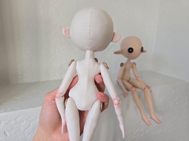 May include: Two handmade doll figures. One doll is held in a hand, showing the back of the doll with a round head, small ears, and jointed limbs. The other doll is seated, with a similar design and a light skin tone.