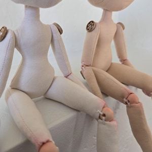 May include: Two handmade cloth dolls with button accents on the shoulders. The dolls are light beige and off-white, with stitched details and articulated limbs. The dolls are sitting, with their legs bent at the knees.