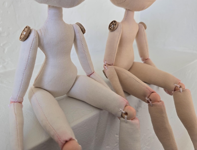 May include: Two handmade cloth dolls with button accents on the shoulders. The dolls are light beige and off-white, with stitched details and articulated limbs. The dolls are sitting, with their legs bent at the knees.
