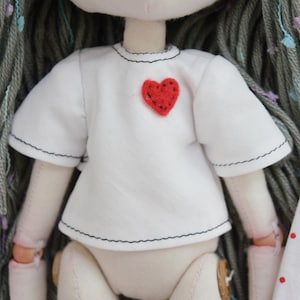 May include: A fabric doll with long gray yarn hair featuring multicolored flecks. The doll wears a short-sleeved white shirt with black stitched edges and a small, textured red heart patch sewn onto the upper left chest area of the garment.