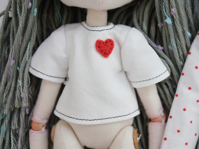 May include: A fabric doll with long gray yarn hair featuring multicolored flecks. The doll wears a short-sleeved white shirt with black stitched edges and a small, textured red heart patch sewn onto the upper left chest area of the garment.