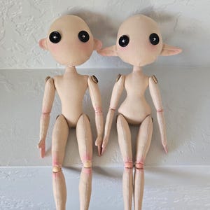 May include: Two handmade cloth dolls with button eyes and rosy cheeks. The dolls have jointed limbs and are made of beige fabric. One doll has small, round ears, while the other has pointed ears. The dolls are sitting side by side.