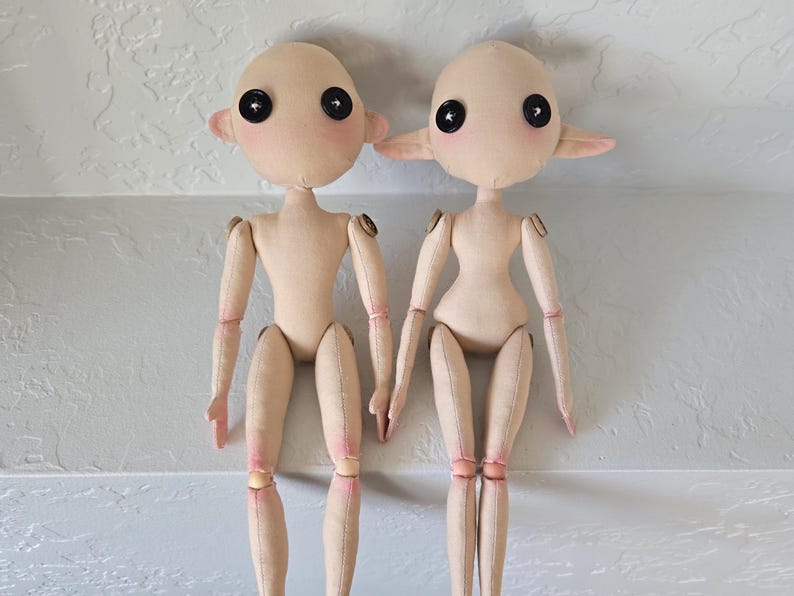 May include: Two handmade cloth dolls with button eyes and rosy cheeks. The dolls have jointed limbs and are made of beige fabric. One doll has small, round ears, while the other has pointed ears. The dolls are sitting side by side.