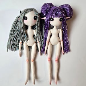 Puede incluir: Dos muñecas de tela hechas a mano con grandes ojos negros y pelo de hilo. Una muñeca tiene el pelo gris, la otra tiene el pelo morado peinado en dos moños. Ambas muñecas tienen la piel pálida, detalles rosas en las extremidades y están de pie.