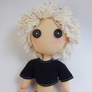 May include: A handmade fabric doll with a beige face, two black button eyes, and messy, textured off-white yarn hair with blue flecks. The doll wears a short-sleeved black crop top with visible white stitching along the hem and sleeve edges.