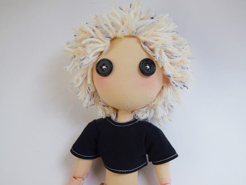 May include: A handmade fabric doll with a beige face, two black button eyes, and messy, textured off-white yarn hair with blue flecks. The doll wears a short-sleeved black crop top with visible white stitching along the hem and sleeve edges.