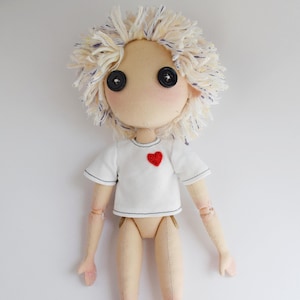 May include: A handcrafted doll with a beige fabric body and articulated joints. The doll features messy, cream-colored yarn hair with blue flecks, two black button eyes, and wears a white short-sleeved t-shirt with a small red heart patch.