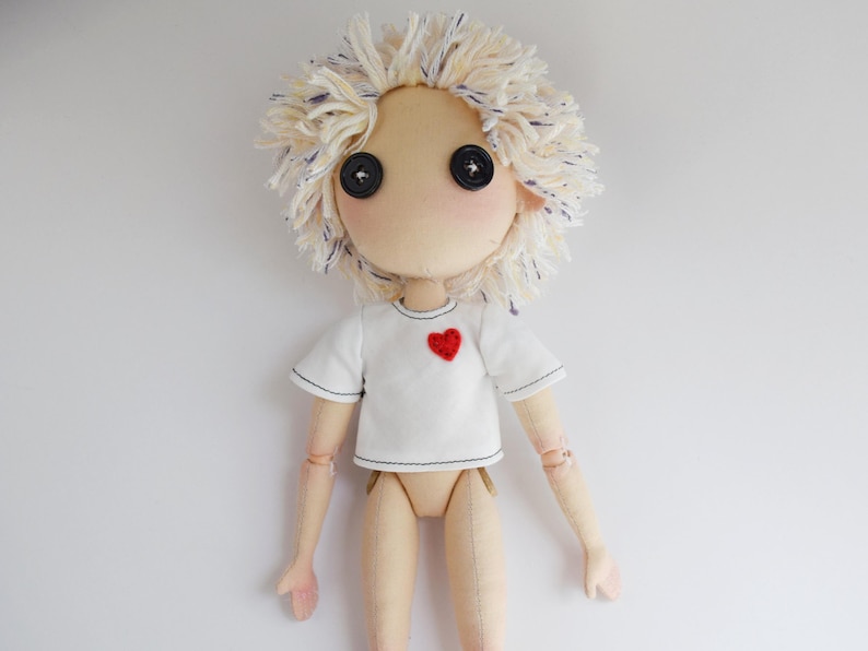 May include: A handcrafted doll with a beige fabric body and articulated joints. The doll features messy, cream-colored yarn hair with blue flecks, two black button eyes, and wears a white short-sleeved t-shirt with a small red heart patch.