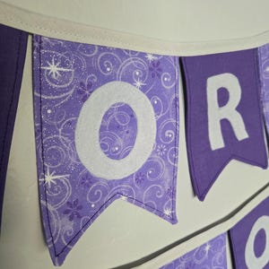 K&ouml;nnte beinhalten: Lila Banner mit wei&szlig;en Buchstaben, die ein Wort bilden. Das Banner hat einen wei&szlig;en Rand und h&auml;ngt an einer wei&szlig;en Wand. Der Buchstabe "O" befindet sich auf einem Banner mit einem Wirbelmuster und der Buchstabe "R" auf einem einfarbigen lila Banner.